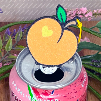 Pink can of sparkling water with a peach Tab Buddy resting on top, surrounded by plants.