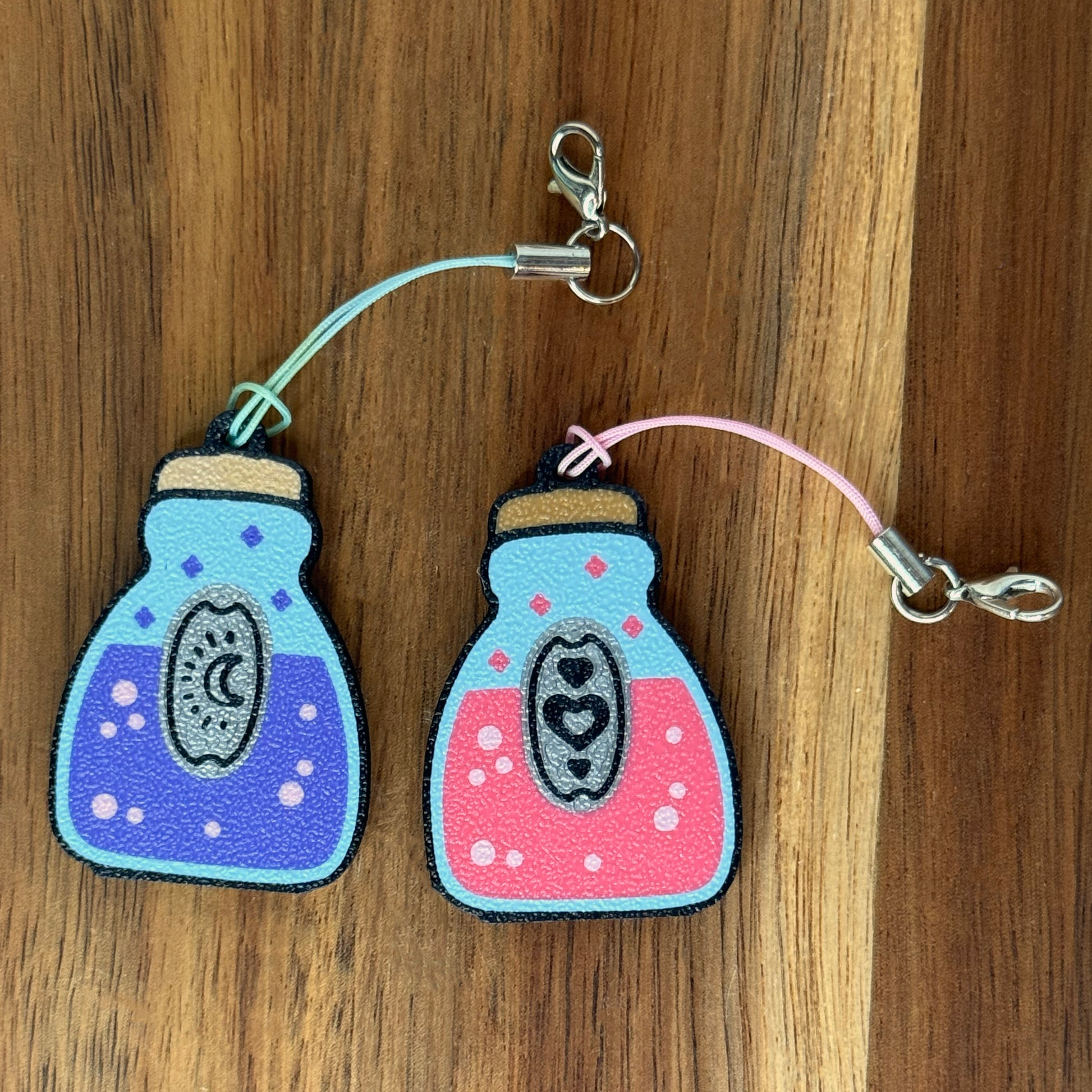 Two potion bottle-shaped Tab Buddy keychains -- one pink and one purple --with colorful contents on a wooden surface