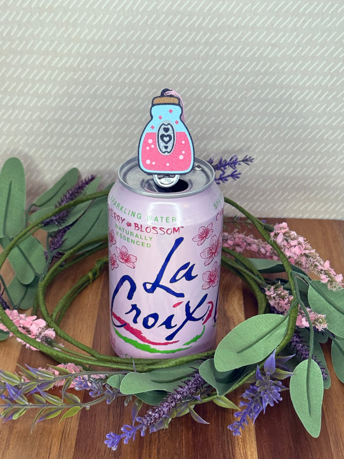 La Croix sparkling water can with a pink potion bottle Tab Buddy resting atop the tab on a wooden surface with greenery and flowers.