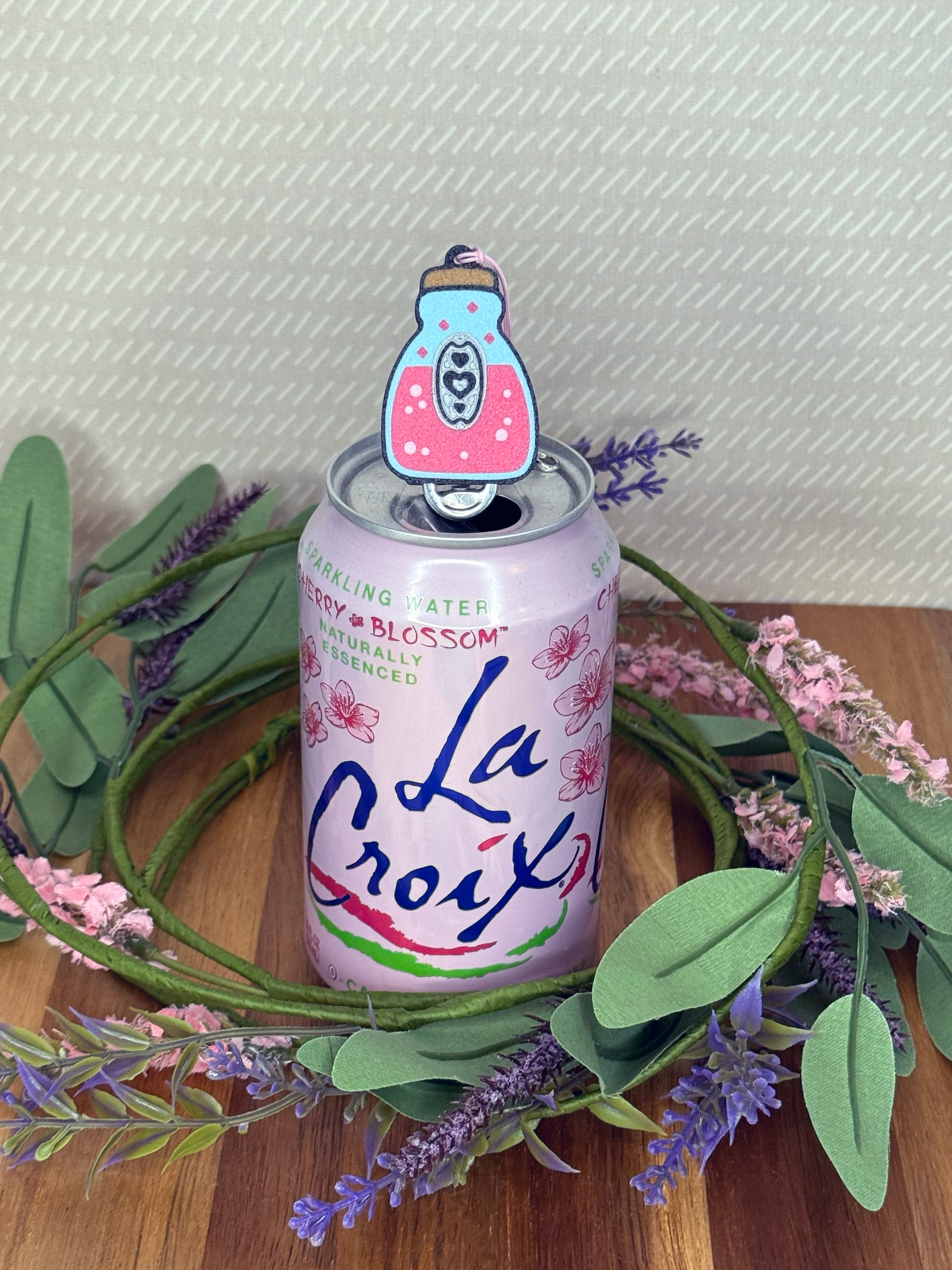 La Croix sparkling water can with a pink potion bottle Tab Buddy resting atop the tab on a wooden surface with greenery and flowers.