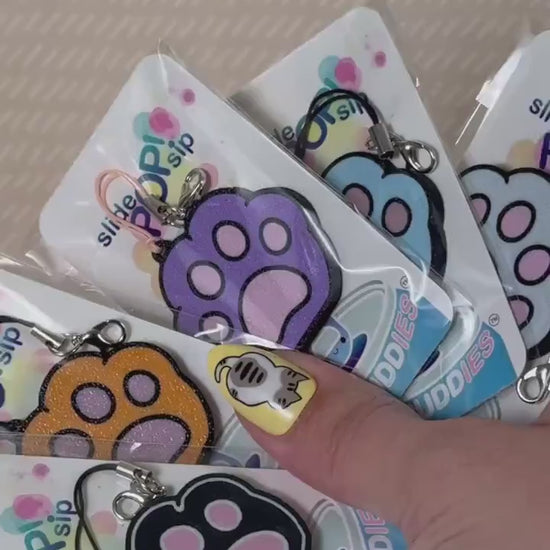 A grey and pink kitty paw Tab Buddy is used to open a red can of Coca-Cola. Other various colored paw Tab Buddies in their packaging also are displayed.