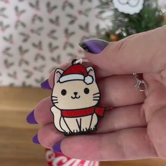 A beige cartoon style cat wearing a red scarf and Santa hat opens a red can of Coca-Cola. Subsequent images show other related Christmas-themed designs of Tab Buddies including a Santa face and a Christmas tree.