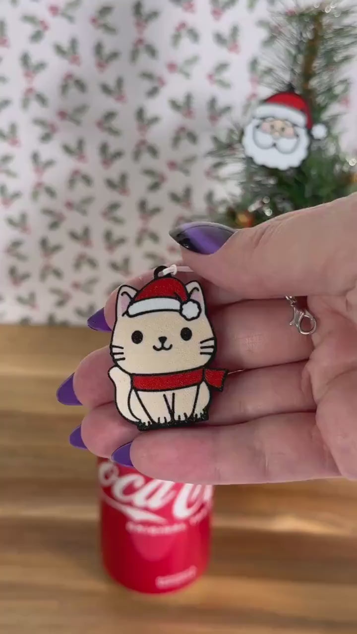 A beige cartoon style cat wearing a red scarf and Santa hat opens a red can of Coca-Cola. Subsequent images show other related Christmas-themed designs of Tab Buddies including a Santa face and a Christmas tree.