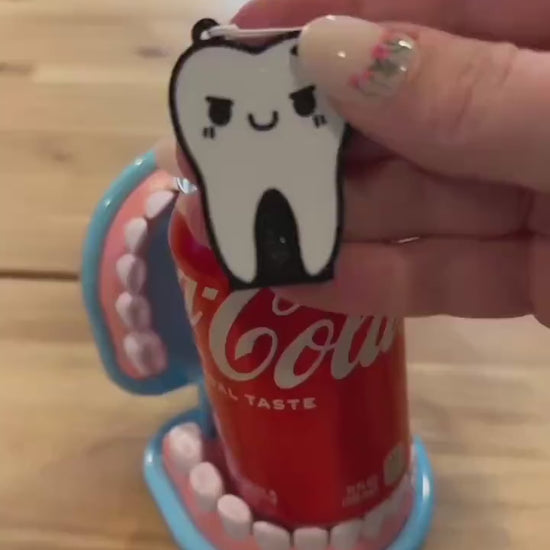 A tooth-shaped Tab Buddy with a mischevious face and crack in it is used to open a red can of Coca-Cola. The can is sitting inside a toy set of dentures.
