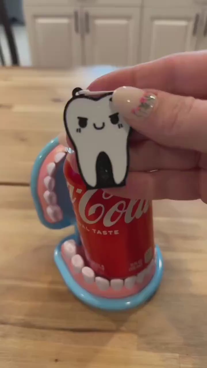 A tooth-shaped Tab Buddy with a mischevious face and crack in it is used to open a red can of Coca-Cola. The can is sitting inside a toy set of dentures.