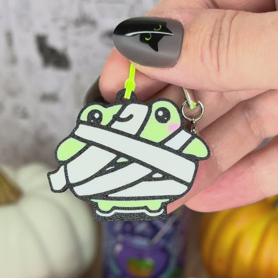 A green Frankenstein monster-themed Halloween frog Tab Buddy is used to open a green Alani can against a backdrop of pumpkins. A mummy frog  Tab Buddy also is shown.
