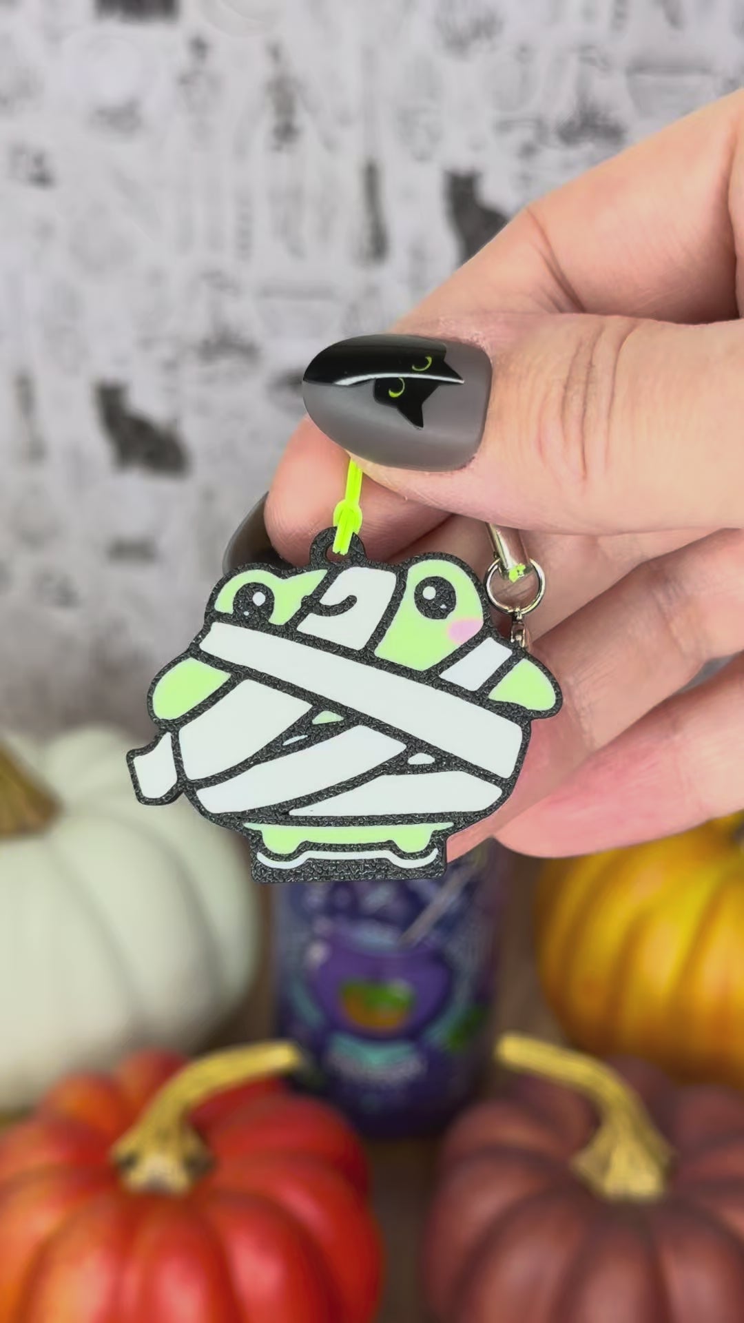 A green Frankenstein monster-themed Halloween frog Tab Buddy is used to open a green Alani can against a backdrop of pumpkins. A mummy frog  Tab Buddy also is shown.