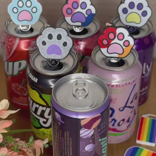 A Kitty Paw Tab Buddy with rainbow colors on the paw pads opens a purple can of Alani. Other colorful paw Tab Buddies rest atop can tabs in the background.