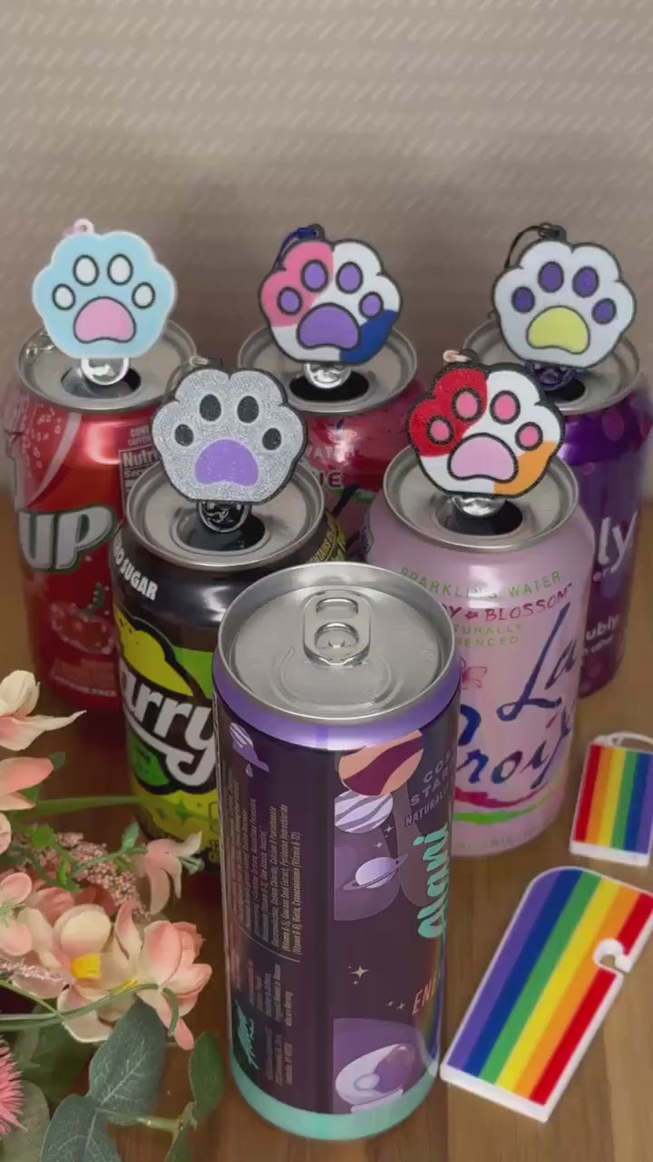 A Kitty Paw Tab Buddy with rainbow colors on the paw pads opens a purple can of Alani. Other colorful paw Tab Buddies rest atop can tabs in the background.