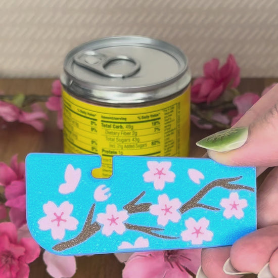 A blue Tab Buddy XL with pink cherry blossom flowers design is used to open a canned food tab. A Great Wave design and Fresh and Fruity design Tab Buddy XL are shown as part of the Spring 2026 lineup.