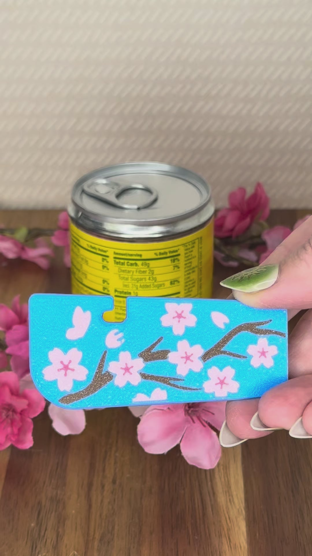 A blue Tab Buddy XL with pink cherry blossom flowers design is used to open a canned food tab. A Great Wave design and Fresh and Fruity design Tab Buddy XL are shown as part of the Spring 2026 lineup.