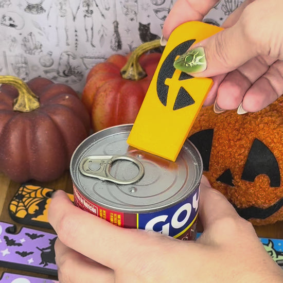 A Jackolantern design Tab Buddy XL opens a can of bean soup by lifting the tab and peeling back its lid. Other Halloween themed designs of Tab Buddy XLs then are displayed.
