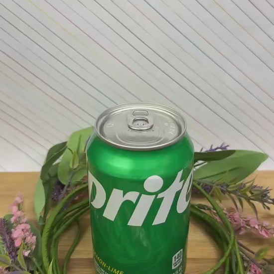 A pink, yellow and beige design Kitty Cone Tab Buddy depicts two cats resting on top of each other as if melting scoops of an ice cream cone. The Tab Buddy is used to open a green can of Sprite sitting on a wooden surface.