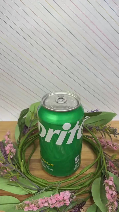 A pink, yellow and beige design Kitty Cone Tab Buddy depicts two cats resting on top of each other as if melting scoops of an ice cream cone. The Tab Buddy is used to open a green can of Sprite sitting on a wooden surface.