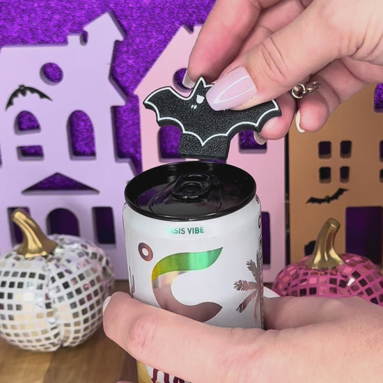 A black bat Tab Buddy keychain is used to open a can of Celsus energy drink against a glittery purple Halloween backdrop. Bats in purple, pink, white and glowing green also are shown against a grey background.
