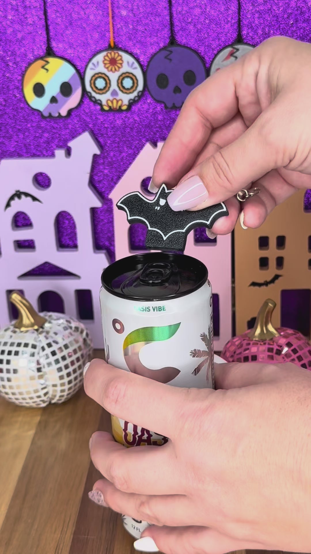 A black bat Tab Buddy keychain is used to open a can of Celsus energy drink against a glittery purple Halloween backdrop. Bats in purple, pink, white and glowing green also are shown against a grey background.
