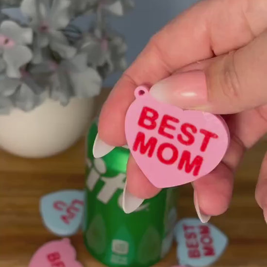 A pink heart Tab Buddy with the phrase "Best Mom" written on it is used to open a green can of Sprite. Other heart Tab Buddies in various colors and phrases surround the can on a wooden surface with a potted flower in the background.