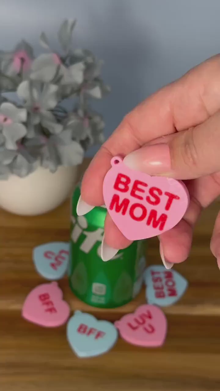 A pink heart Tab Buddy with the phrase "Best Mom" written on it is used to open a green can of Sprite. Other heart Tab Buddies in various colors and phrases surround the can on a wooden surface with a potted flower in the background.