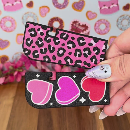 A black Tab Buddy XL with hearts design is used to lift the tab on a can of soup, then peel back the lid. Four color options of each the Hearts design XL and a Leopard print design XL are shown.