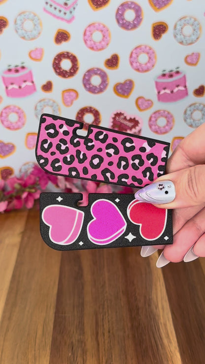 A black Tab Buddy XL with hearts design is used to lift the tab on a can of soup, then peel back the lid. Four color options of each the Hearts design XL and a Leopard print design XL are shown.