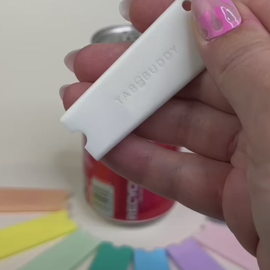 A white Tab Buddy Classic is used to open a red can of Coca-Cola. Other colorful Tab Buddy Classics surround the can.