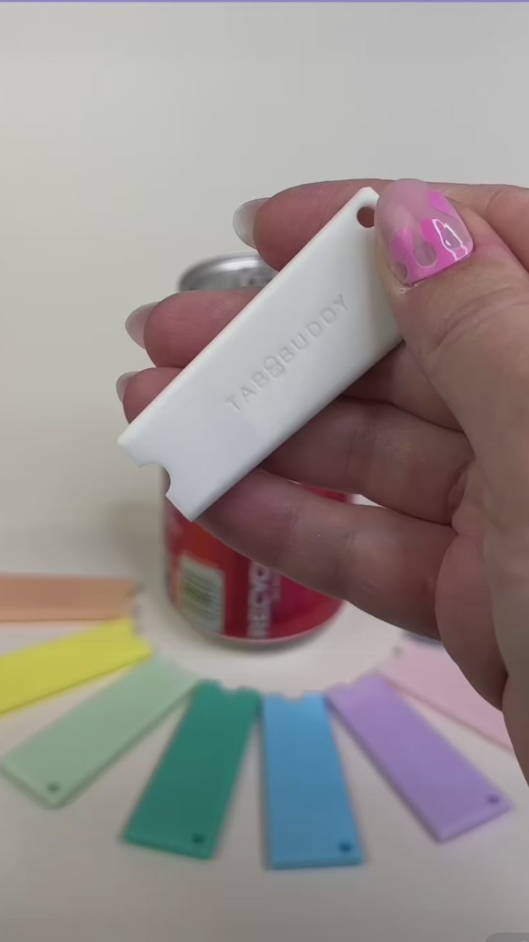 A white Tab Buddy Classic is used to open a red can of Coca-Cola. Other colorful Tab Buddy Classics surround the can.
