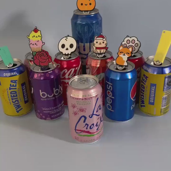 A Tab Buddy designed to look like a Gingerbread Cat Cookie is used to open a pink can of La Croix. A variety of other  cans rest in the background with colorful Tab Buddies resting atop their tabs.