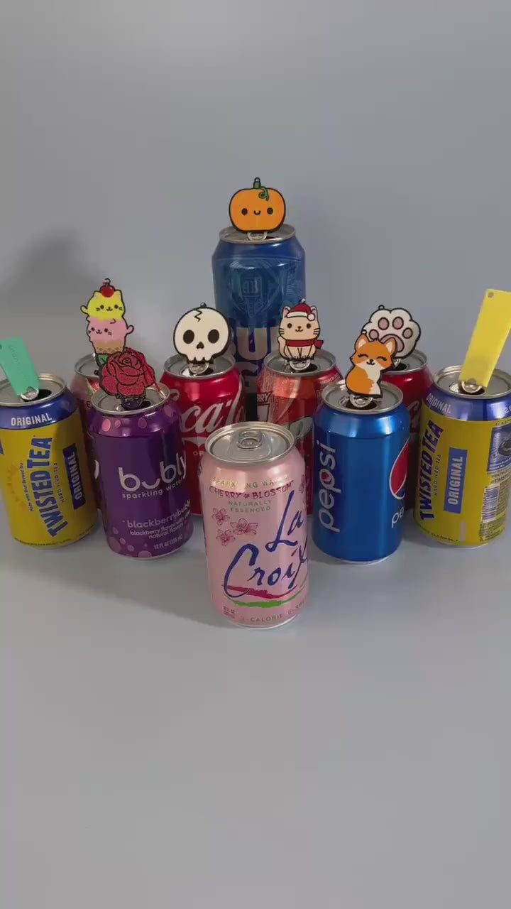 A Tab Buddy designed to look like a Gingerbread Cat Cookie is used to open a pink can of La Croix. A variety of other  cans rest in the background with colorful Tab Buddies resting atop their tabs.