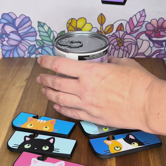 A Tab Buddy XL with a black-and-white cat and golden eyes design is used to open a can of pet food. Other Tab Buddy XLs with different color cats surround the can that rests on a wooden surface with floral backdrop.