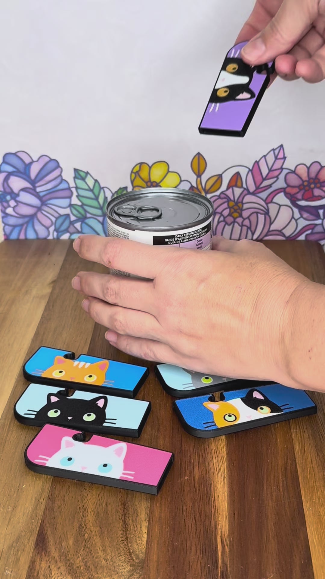 A Tab Buddy XL with a black-and-white cat and golden eyes design is used to open a can of pet food. Other Tab Buddy XLs with different color cats surround the can that rests on a wooden surface with floral backdrop.