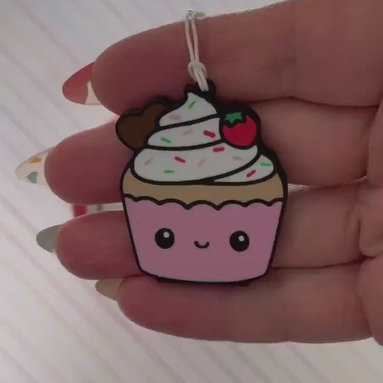 A Tab Buddy designed to be a pink-and-white cupcake with sprinkes and a strawberry on top is used to open a red can of Coca-Cola. The can rests atop a background of diagonal rainbow stripes.