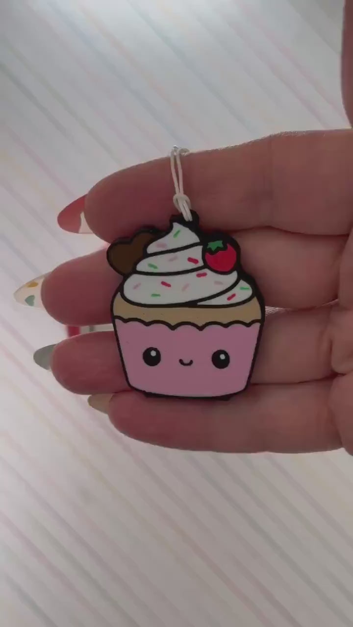 A Tab Buddy designed to be a pink-and-white cupcake with sprinkes and a strawberry on top is used to open a red can of Coca-Cola. The can rests atop a background of diagonal rainbow stripes.