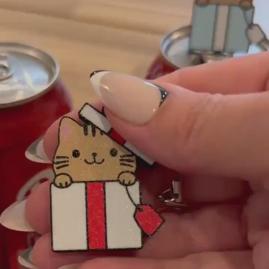 a light brown kitty peeking its head out of a red-and-white giftbox is the design on a Tab Buddy that opens a red can of Coca-Cola sitting on a wood table. Another Tab Buddy of the same design but with a blue and white giftbox sits atop a neighboring can.