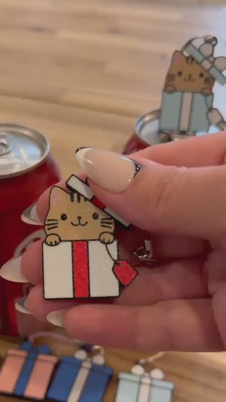 a light brown kitty peeking its head out of a red-and-white giftbox is the design on a Tab Buddy that opens a red can of Coca-Cola sitting on a wood table. Another Tab Buddy of the same design but with a blue and white giftbox sits atop a neighboring can.