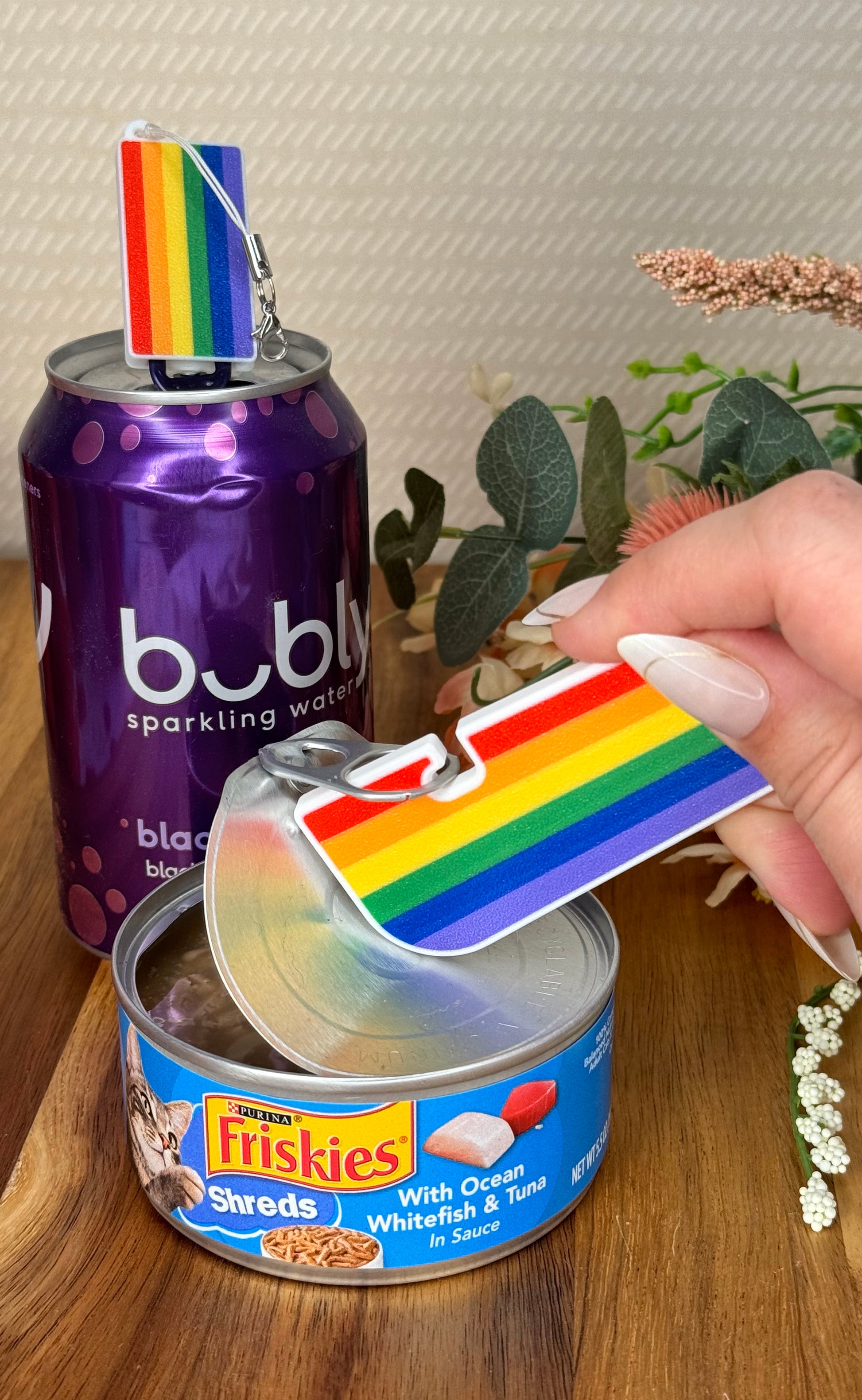 Hand holding a can of Friskies cat food with a rainbow flag Tab Buddy XL peelingback the lid, next to a can of Bubly sparkling water with a matching soda-sized Tab Buddy.