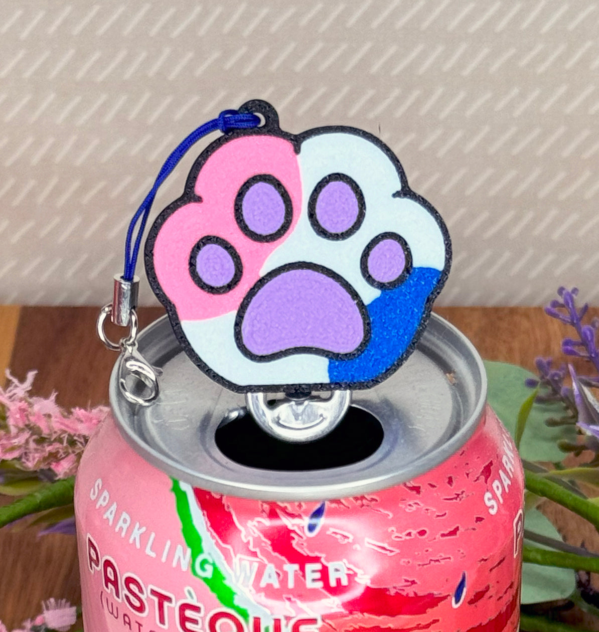 Pink can of sparkling water with a colorful pink, purple, white and blue paw-shaped Tab Buddy keychain on a wooden surface.