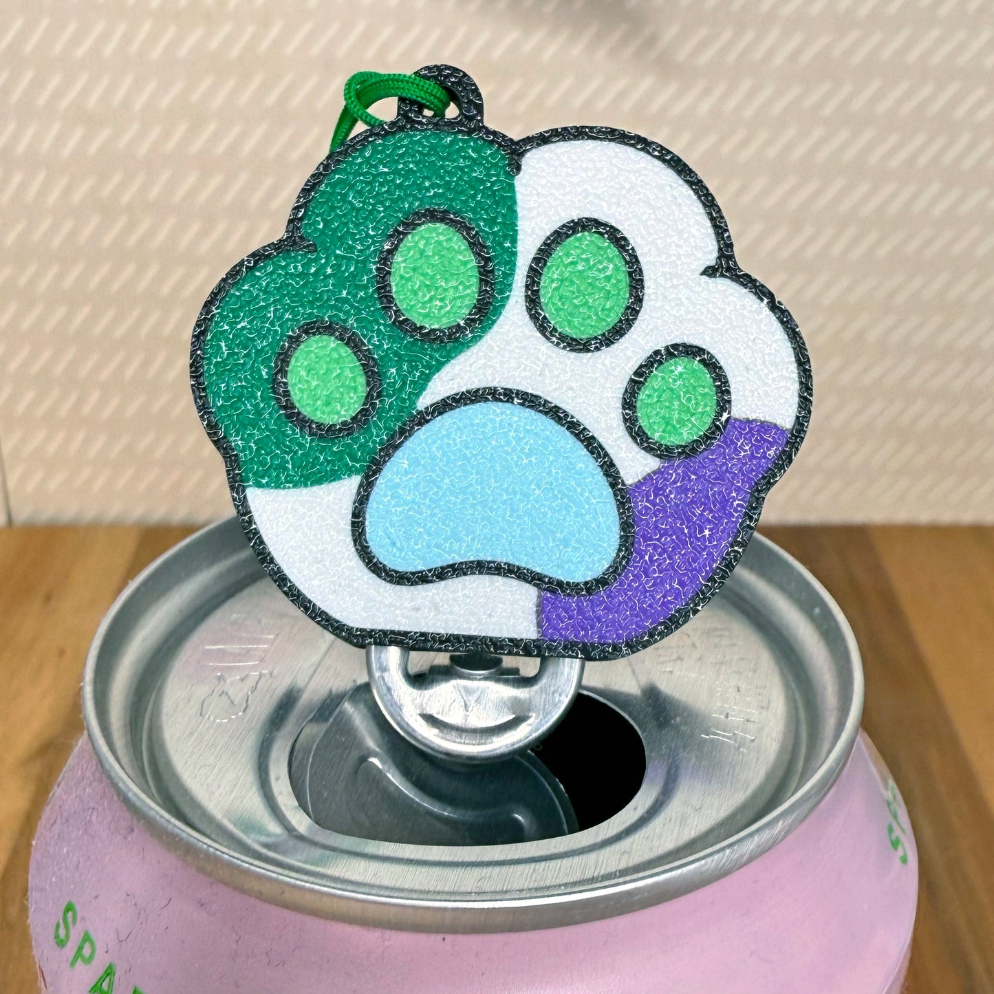 Colorful green, blue and purple Pride paw print Tab Buddy on a can opener sits atop a pink can