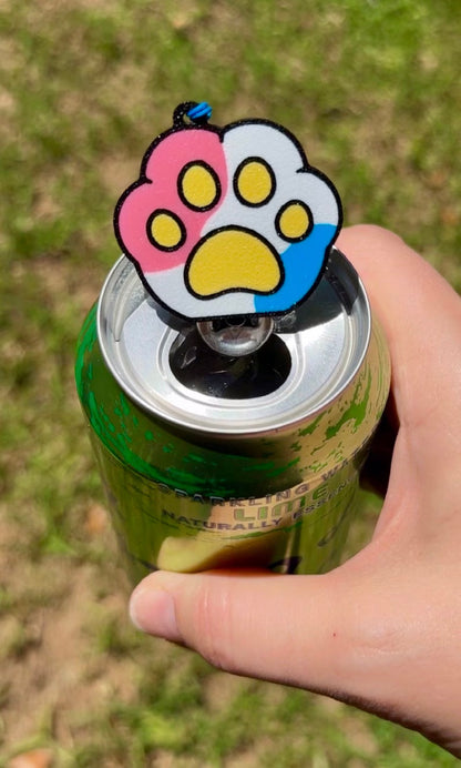 Hand holding a can with a pink, white, yellow and blue paw-shaped Tab Buddy on a grassy background