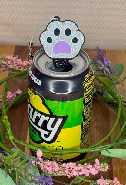 Can of soda with a paw-shaped silver, black and purple Tab Buddy keychain on a wooden surface with flowers.