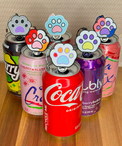 Collection of soda cans with paw-shaped Tab Buddies on a wooden surface.