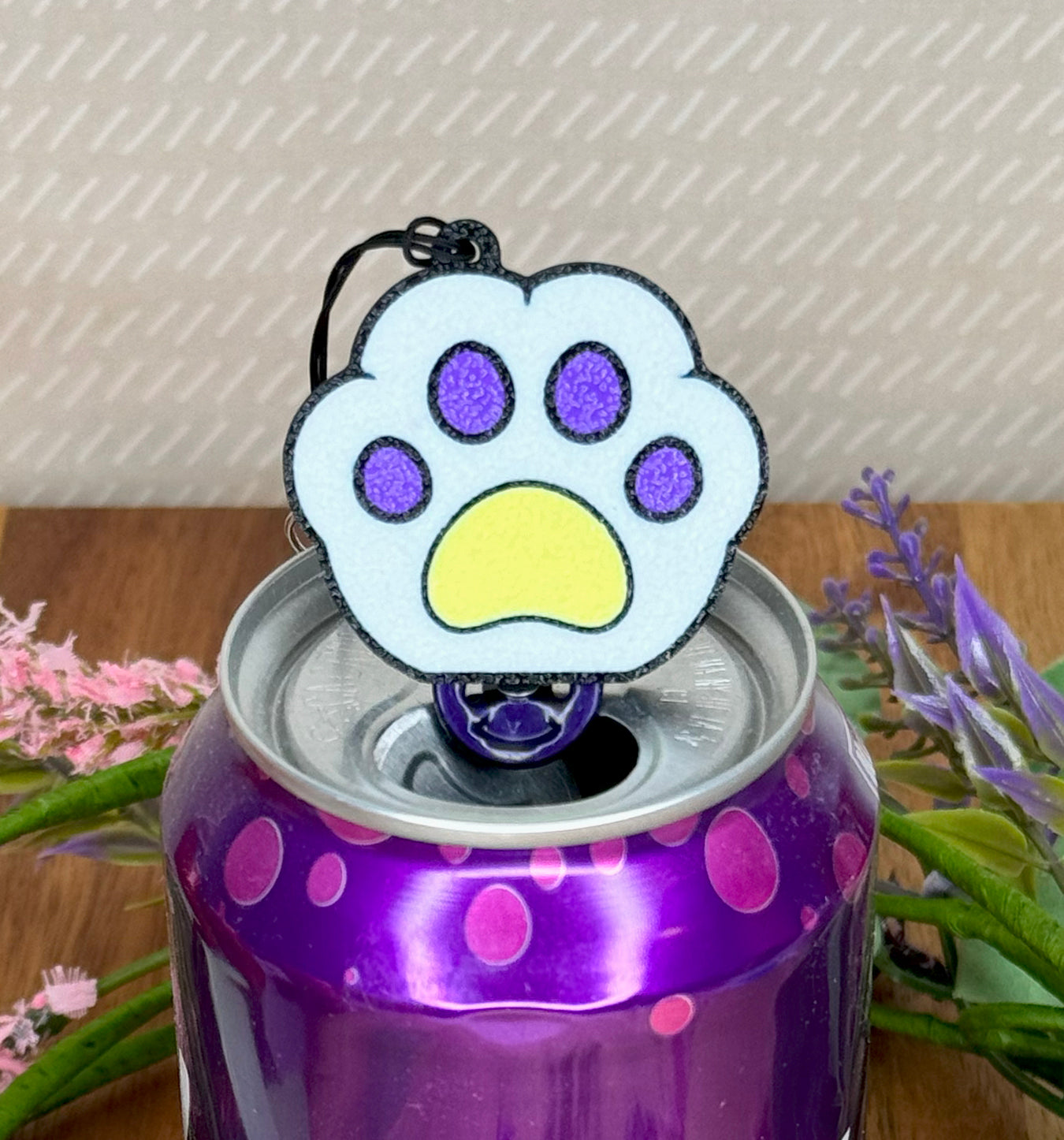 Purple soda can with a white, purple and yellow paw print Tab Buddy on top, surrounded by flowers.