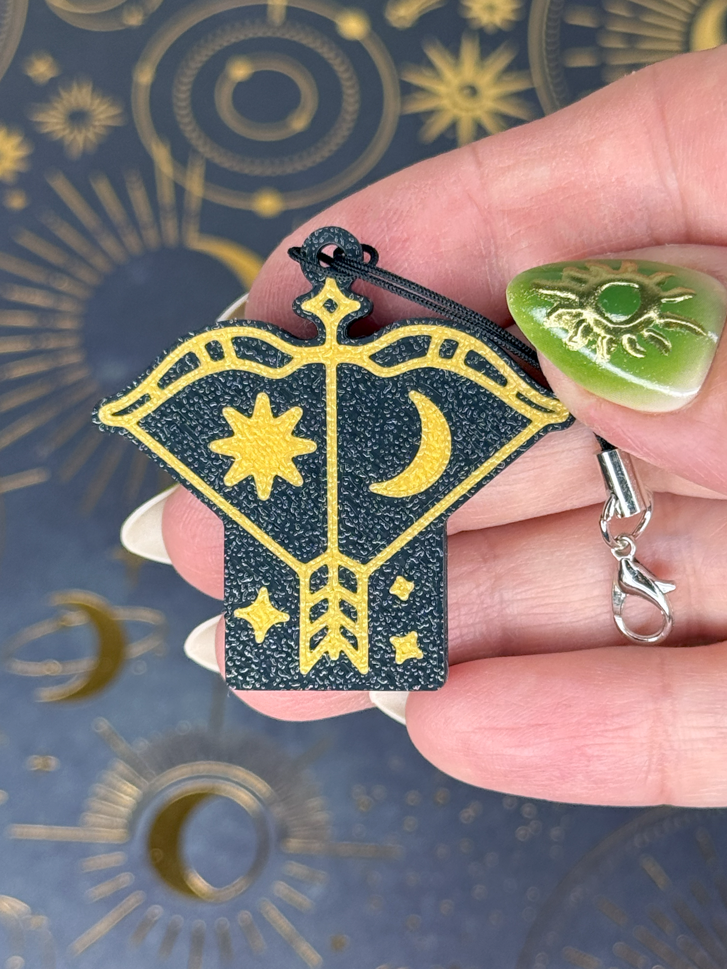 Hand holding a black and gold Sagittarius Tab Buddy with star and moon design against a patterned background