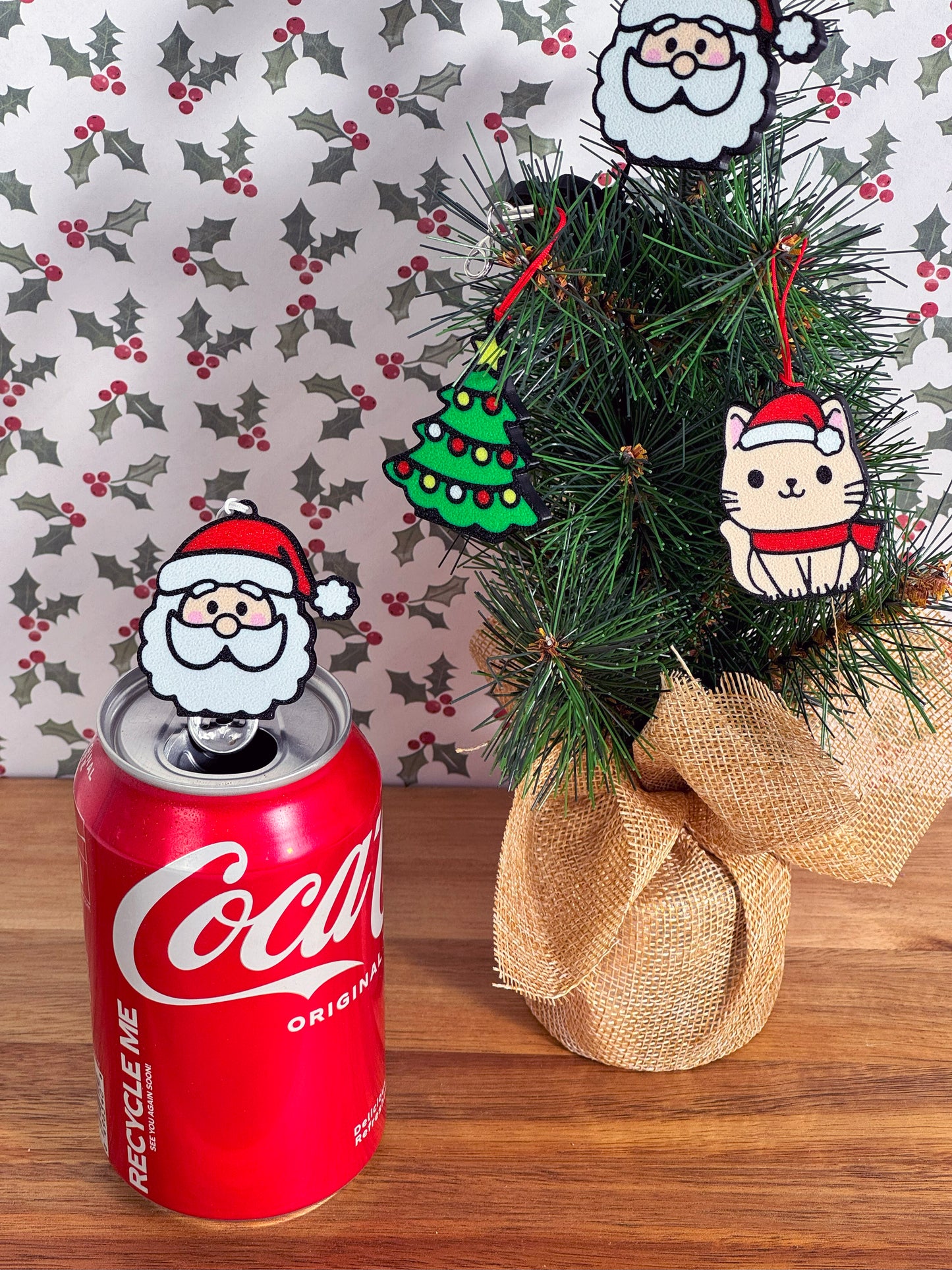 Coca-Cola can with a Santa head-shaped Tab Buddy resting on top next to a small Christmas tree with more Tab Buddies hanging as ornaments and a patterned background