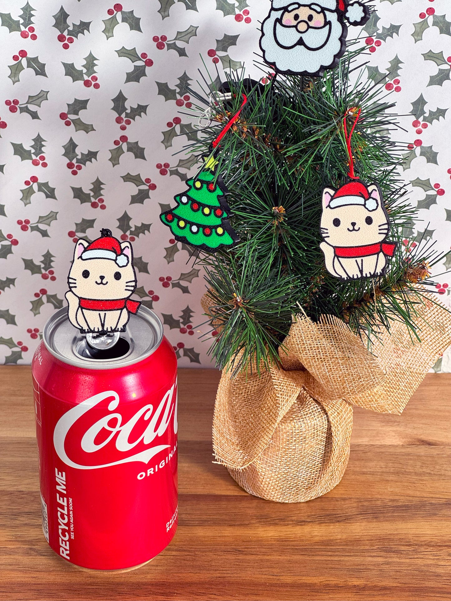 Coca-Cola can with a beige cat wearing a Santa hat atop the tab next to a small decorated Christmas tree with more Tab Buddies hanging as ornaments on a wooden surface.