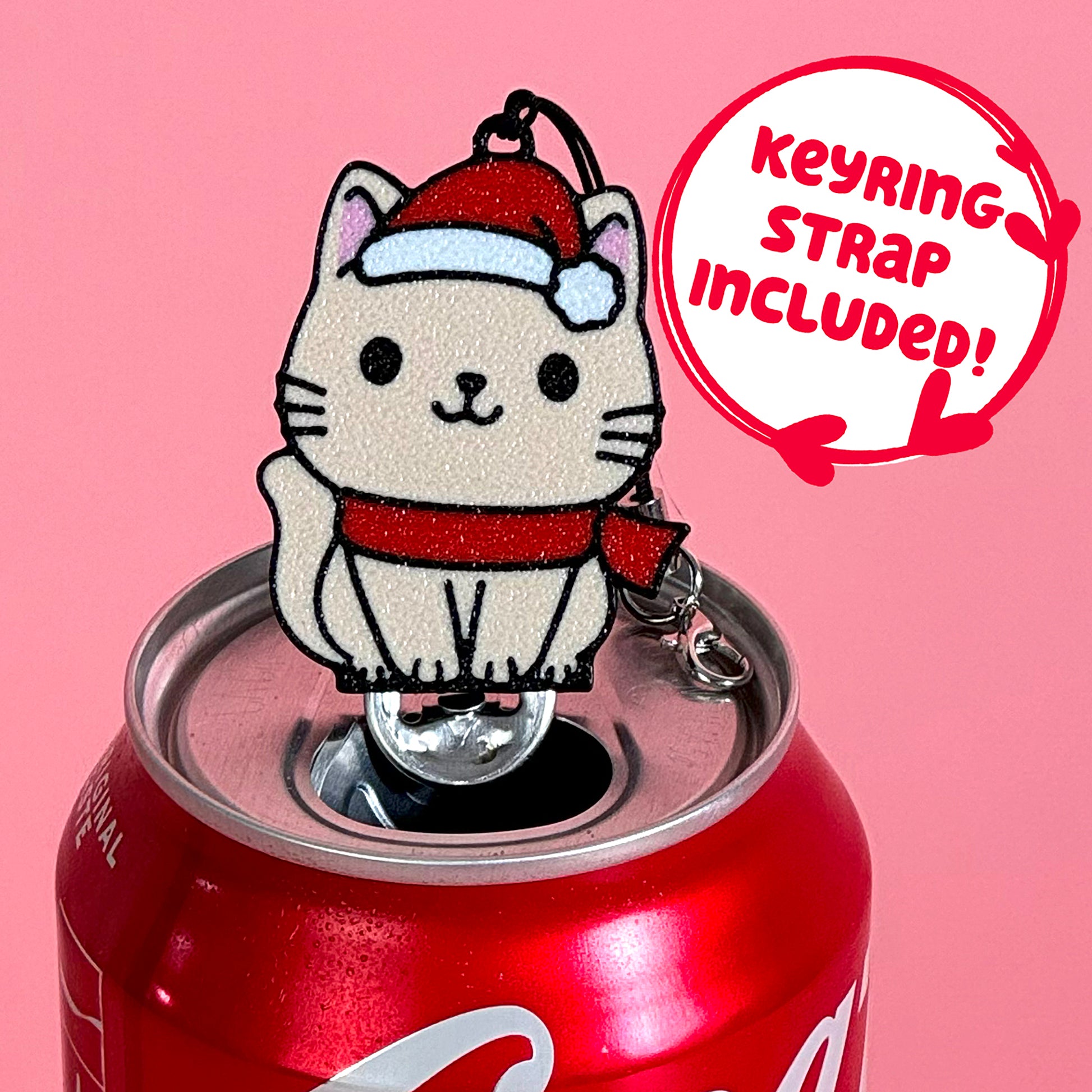 Santa cat Tab Buddy keychain sits atop a Coca-Cola can against a pink background