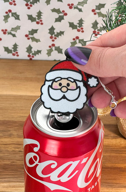 Coca-Cola can with a Santa Claus Tab Buddy on top on a wooden surface with a holly leaf pattern in the background.