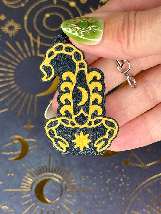 Decorative Tab Buddy keychain with gold and black Zodiac Scorpio design held by a hand against a celestial patterned background.
