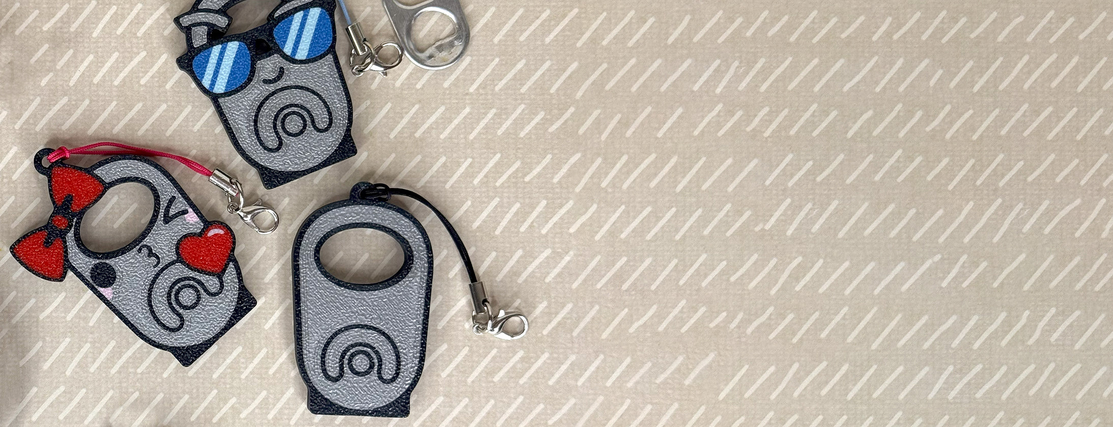 Three soda tab themed Tab Buddy keychains are shown atop a textured beige background