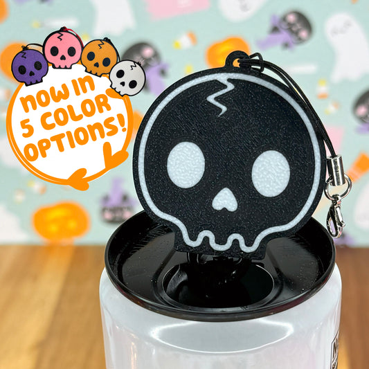 Black skull-shaped Tab Buddy keychain sits atop a black and white soda can tab with Halloween-themed background. Text reads "Now in 5 color options."