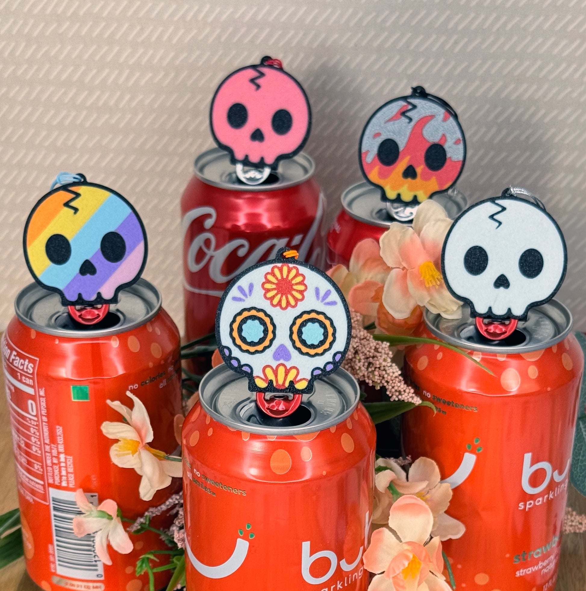 Various soda cans with colorful skull-shaped Tab Buddies on top on a textured surface.